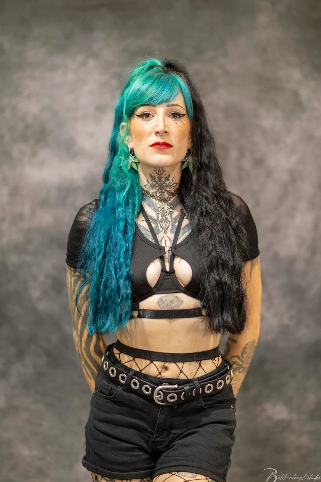 Miss CBD en studio, cheveux mi-bleu mi-noir, tatouée, en tenue noire